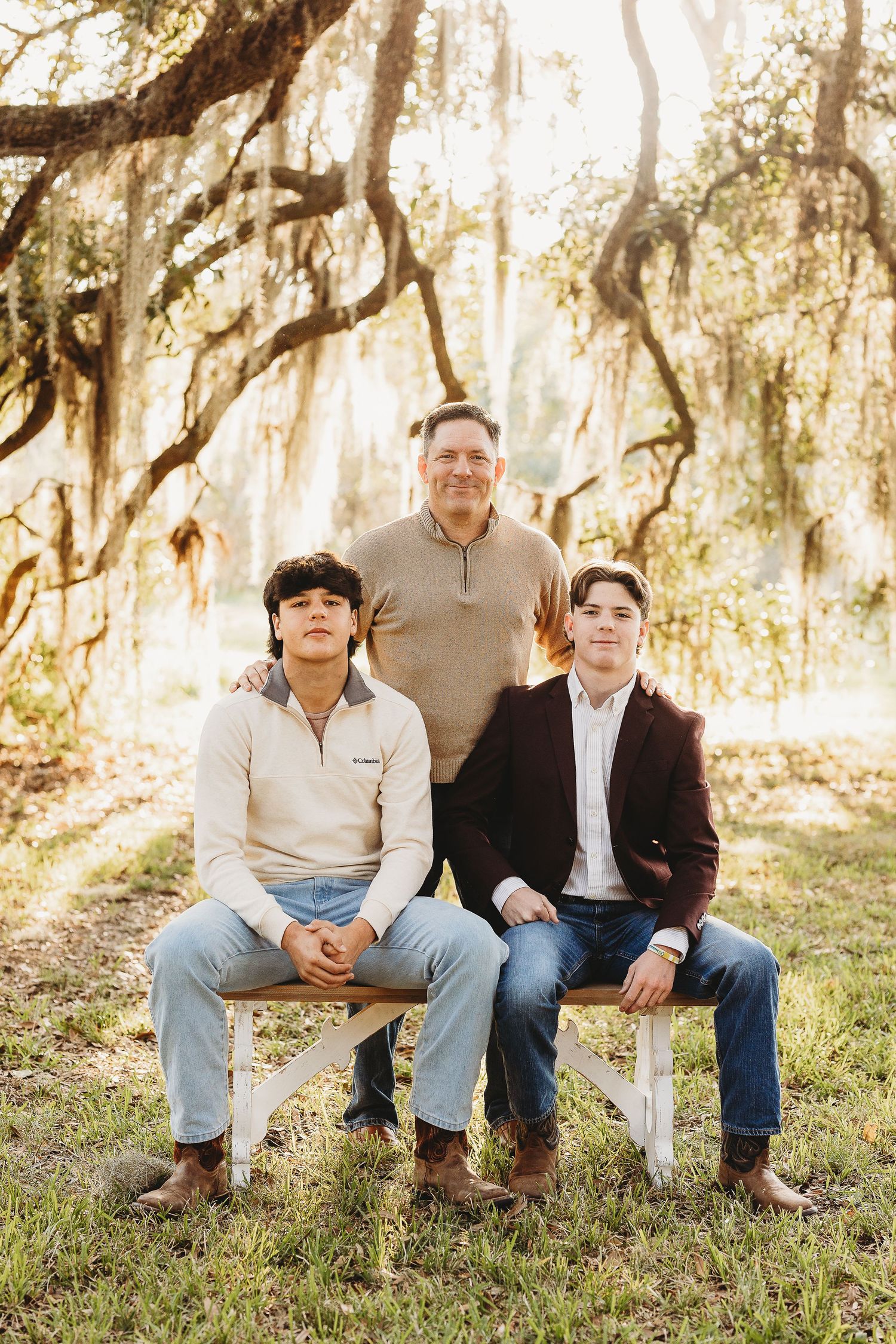 Three people pose together on wooden benches under Spanish moss-draped trees in a sunlit outdoor setting.