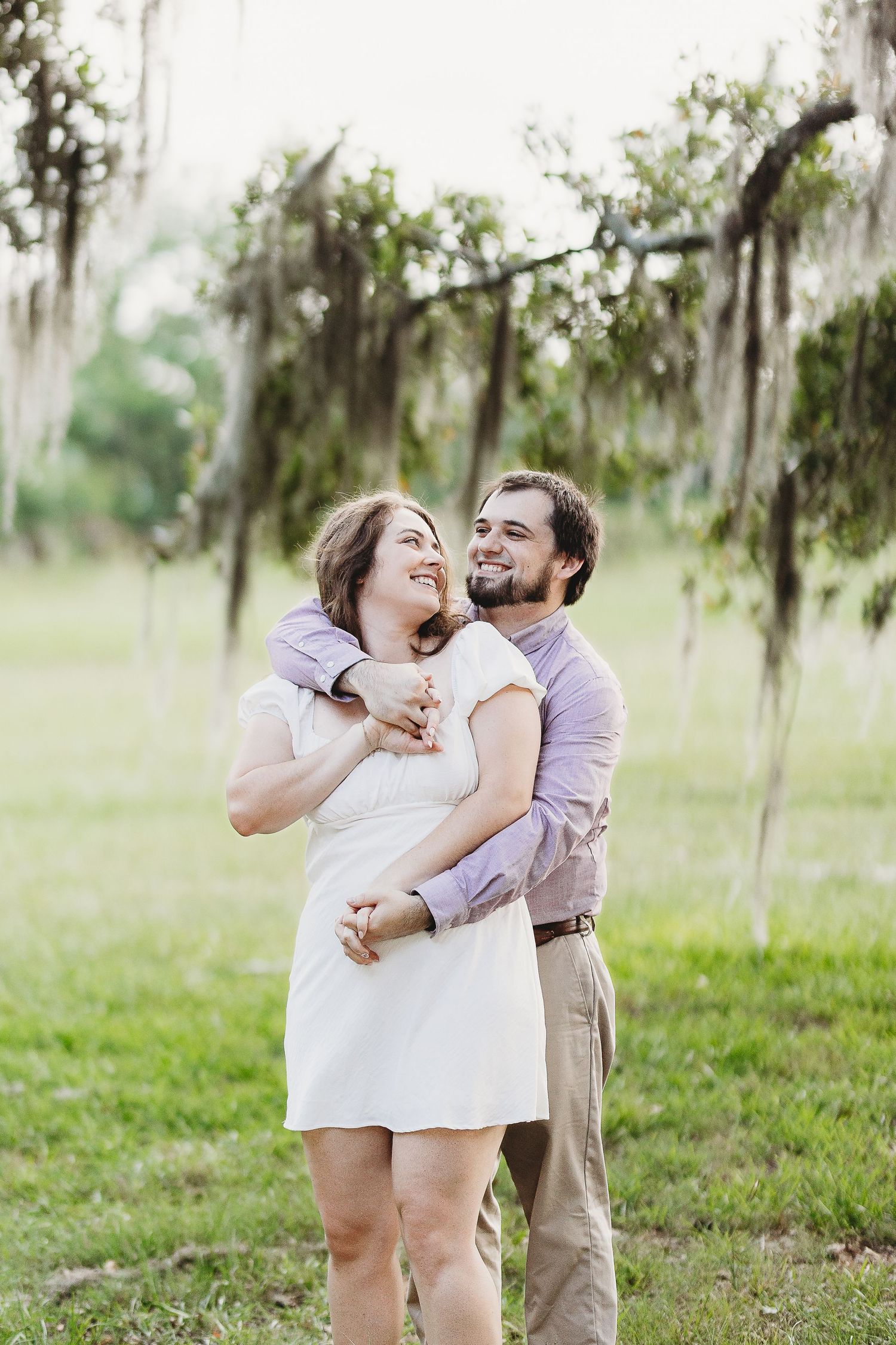A playful couple embraces in a grassy field with Spanish moss hanging from trees.