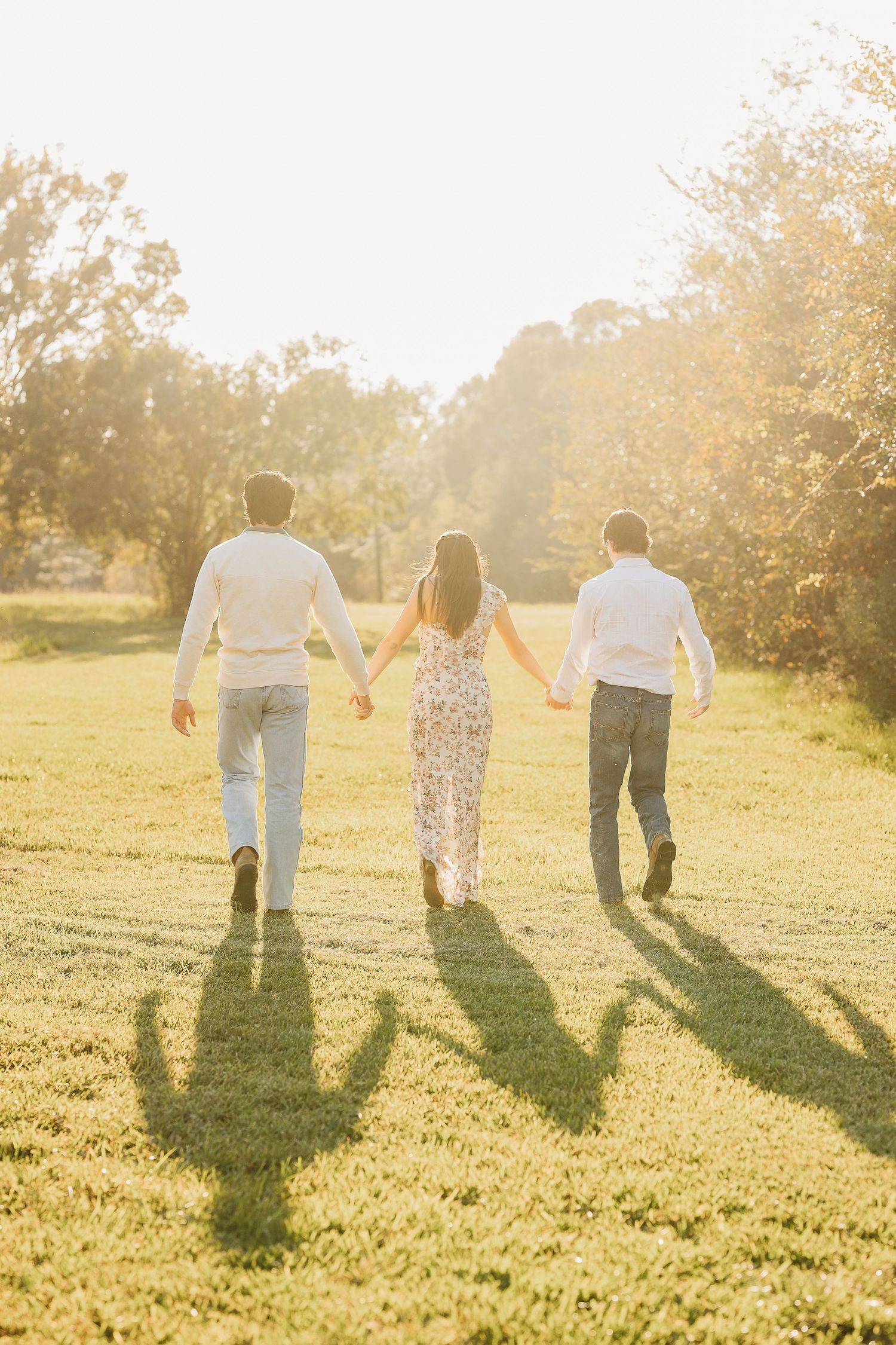 Three people walk hand in hand into the sunset casting long shadows on grassy field.