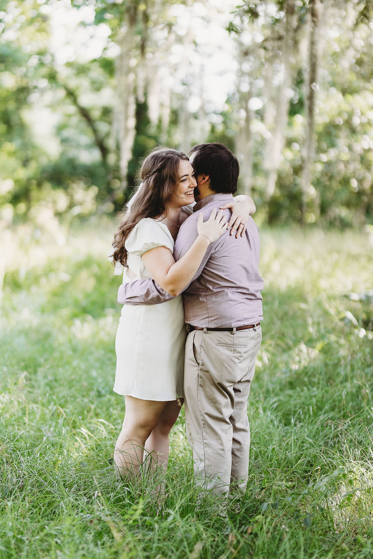 A couple shares tender moments while standing in tall grass under moss-draped trees.