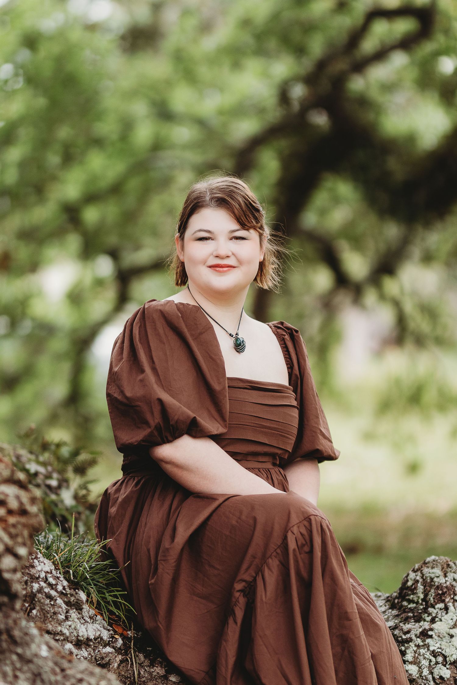 A portrait of someone in a brown vintage-style dress sitting on mossy rocks.