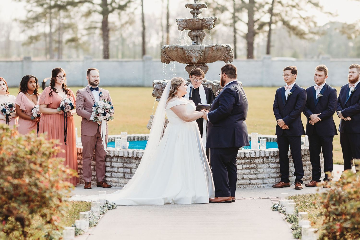 Wedding ceremony takes place outdoors with tall pine trees and a decorative stone fountain in the background.