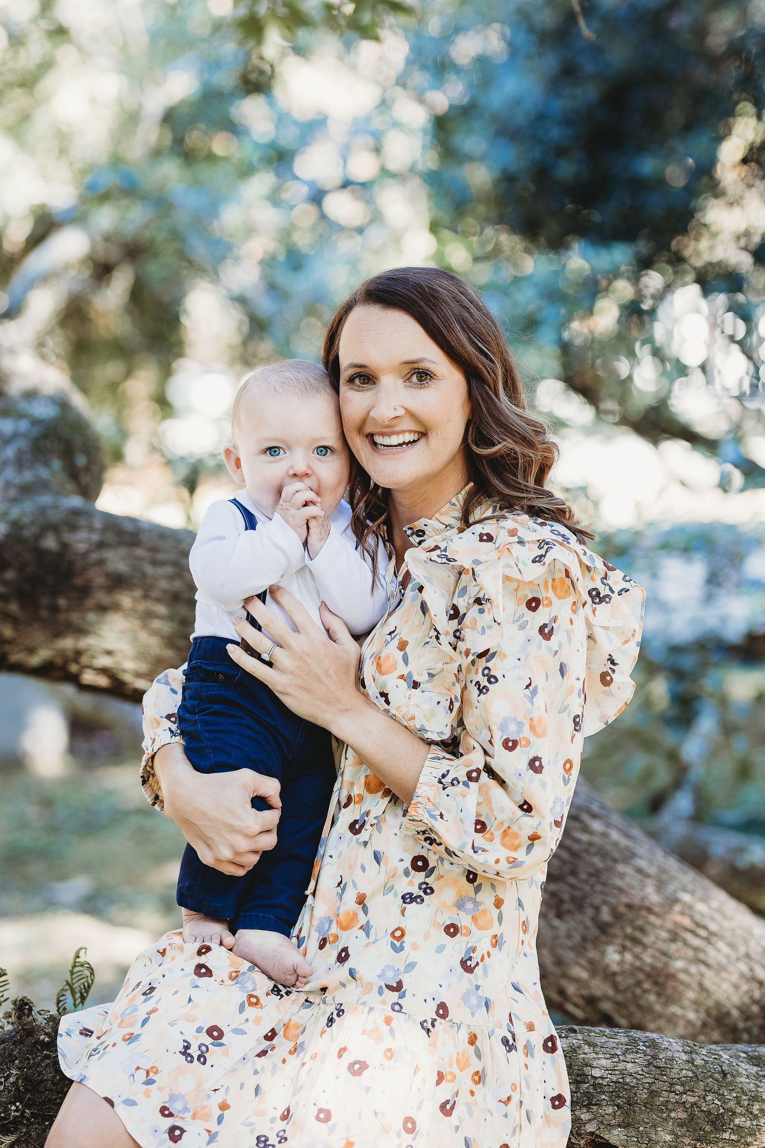 A floral dress-wearing woman holds a baby in blue against a dreamy outdoor backdrop with bokeh lighting effects.