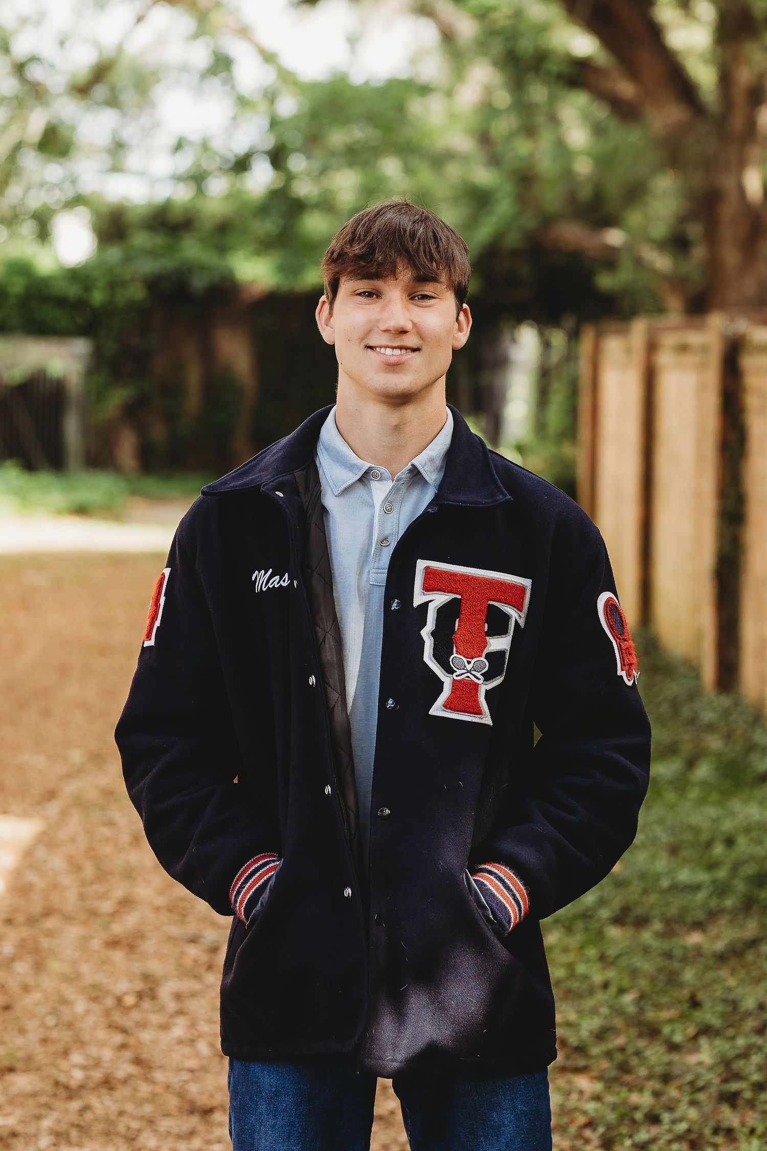 Student wearing varsity letterman jacket with T emblem stands on outdoor path.
