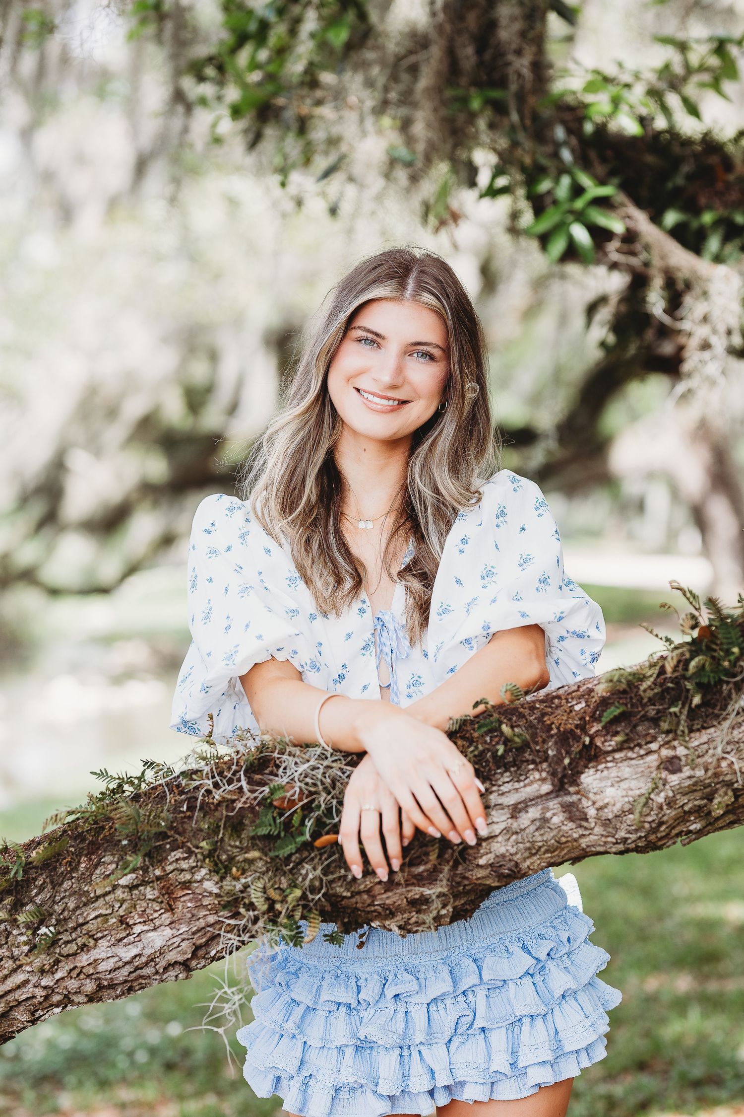 Portrait session featuring floral top and denim styling with natural moss-draped trees in background.