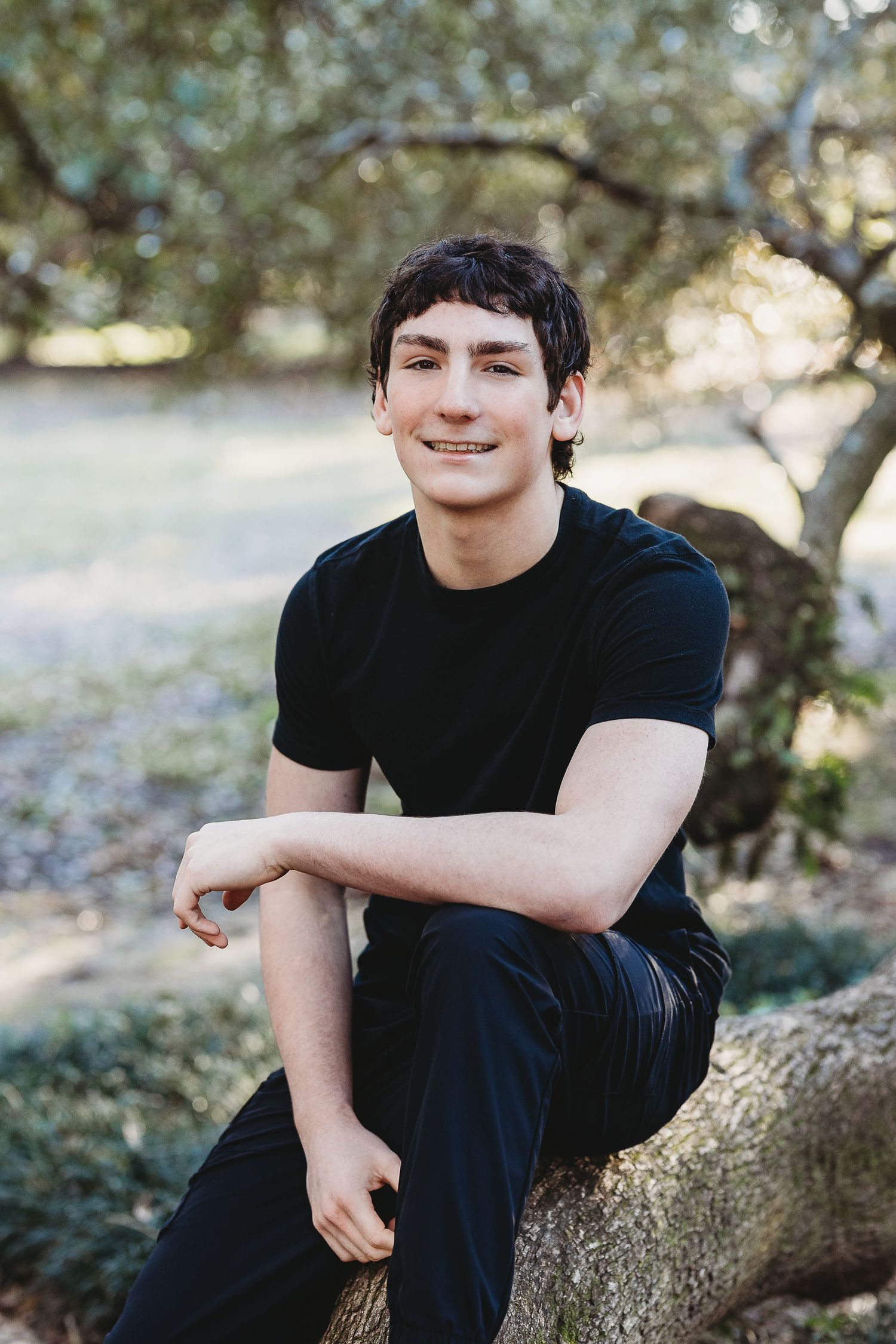 Student in black shirt sitting casually on tree trunk in outdoor setting.