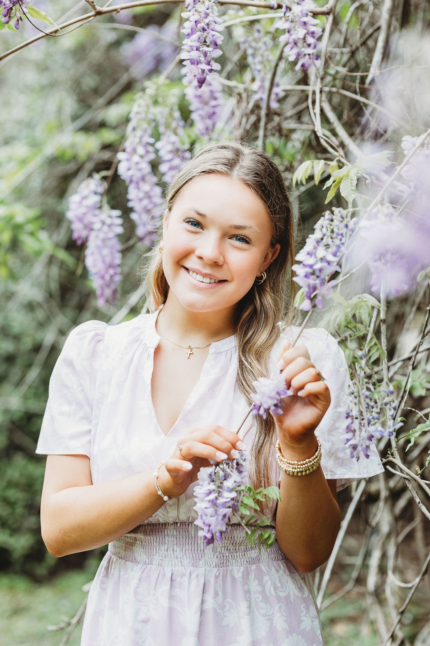 Someone in a white dress poses with blooming purple wisteria flowers in a garden setting.