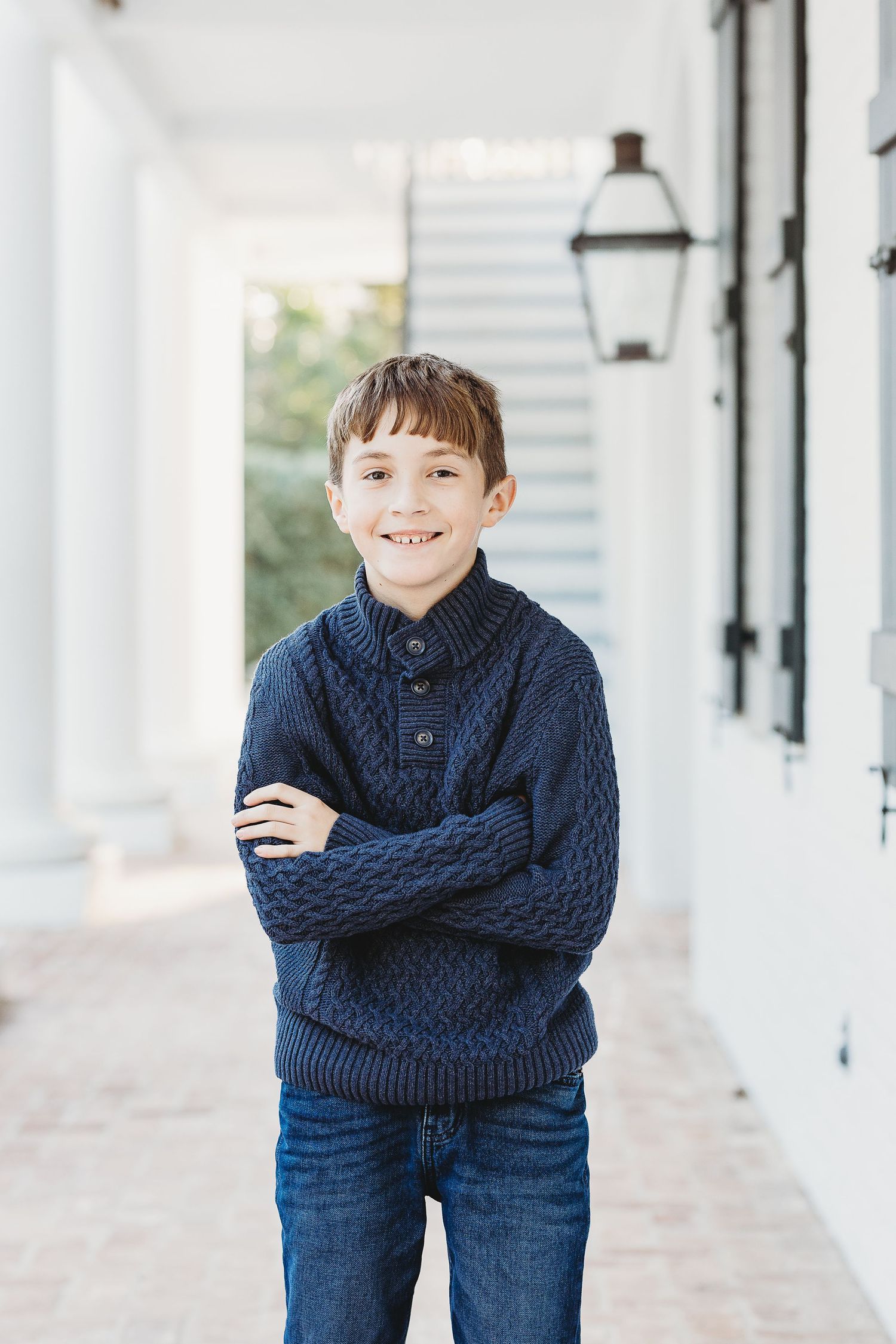 Young person in navy cable knit sweater and jeans poses playfully in white columned hallway.