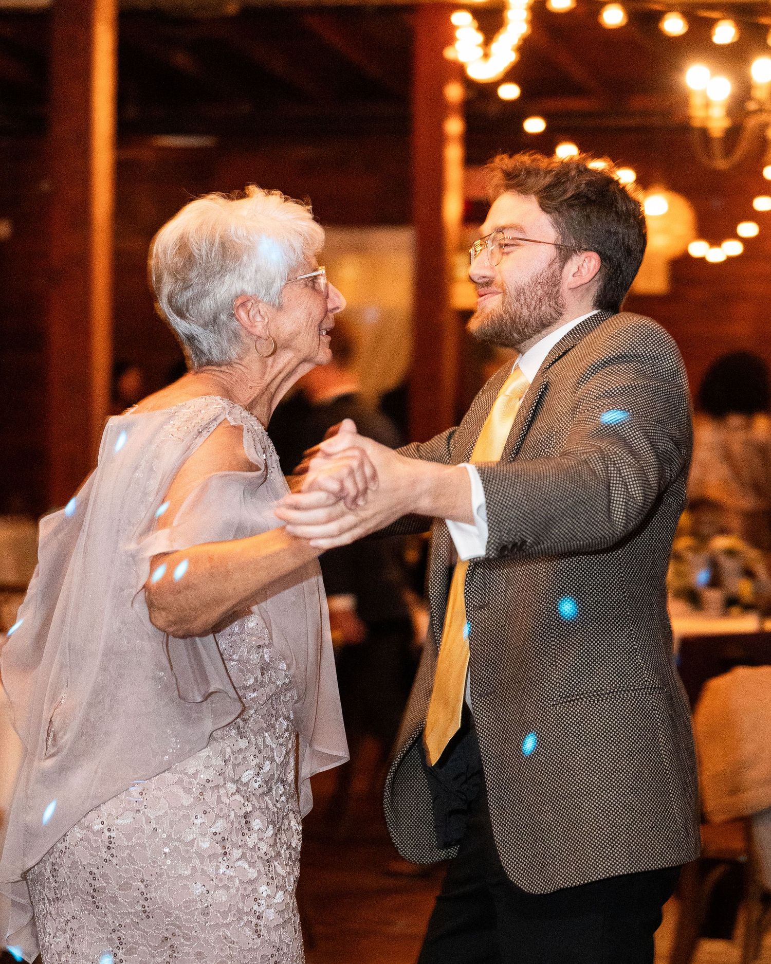 A joyful dance moment between two people at an indoor reception with warm lighting and string lights overhead.