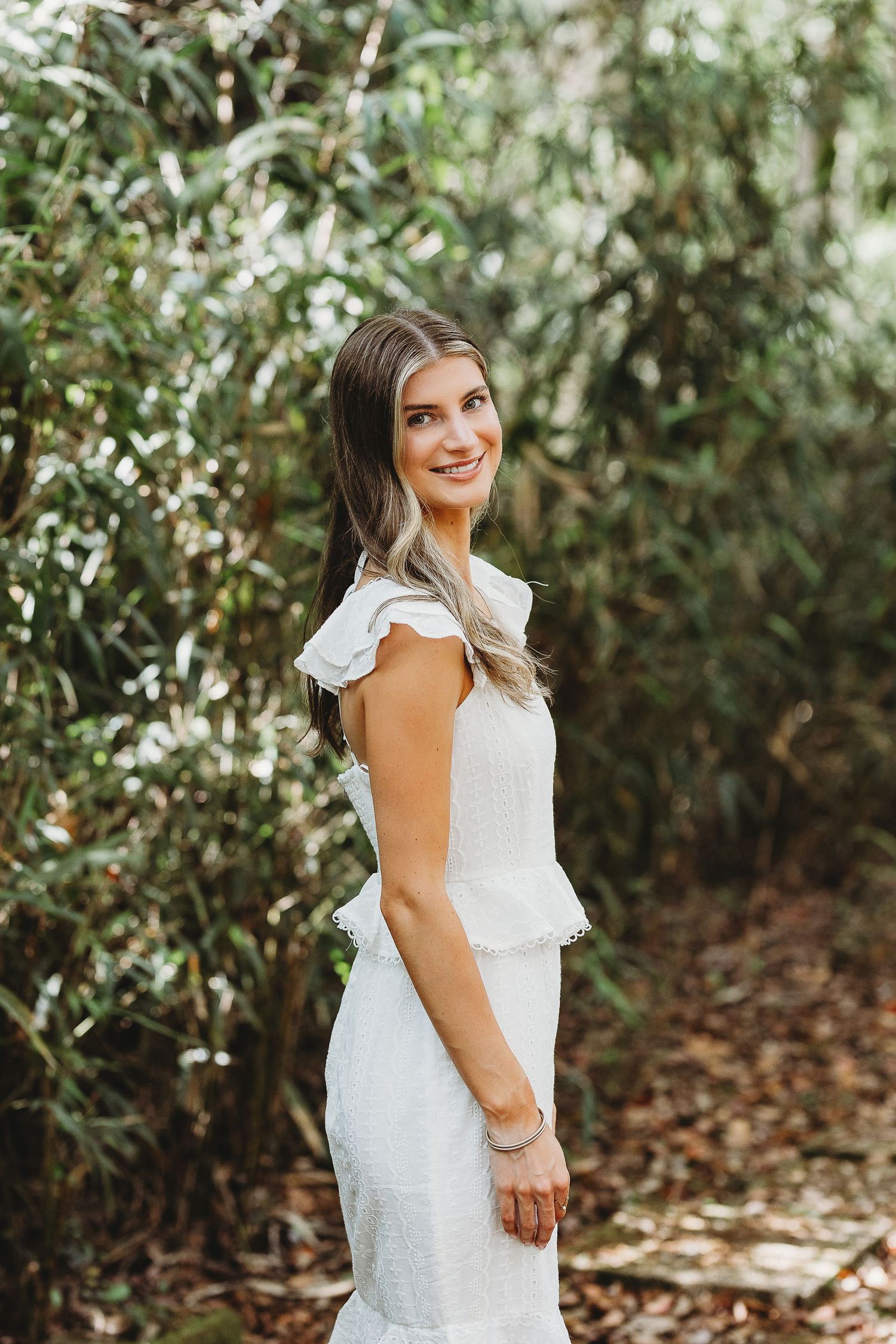 A woman in a white ruffled dress poses outdoors against a natural green foliage background.