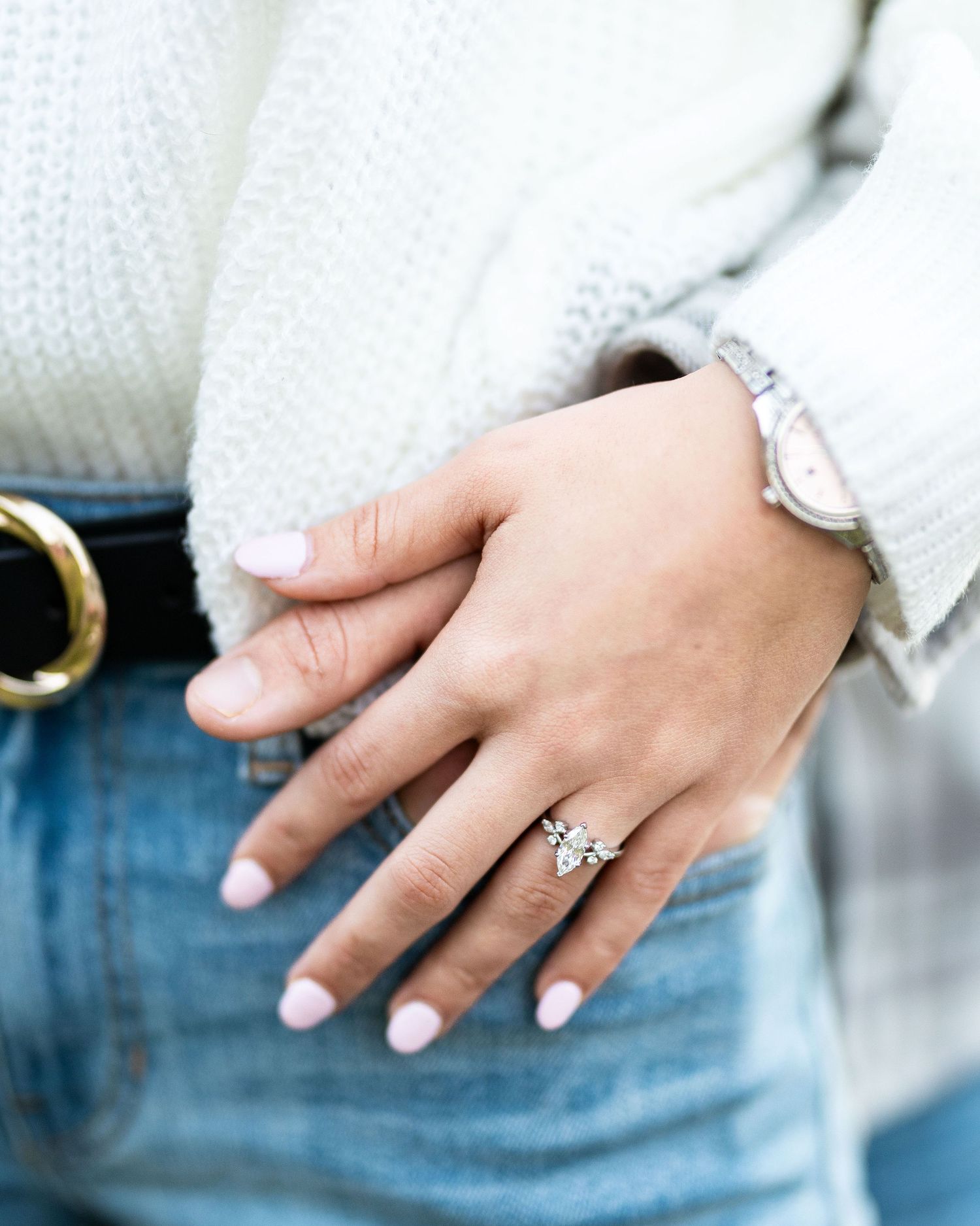 Close-up of a sparkling diamond engagement ring on a woman's hand with denim and white sweater in the background.