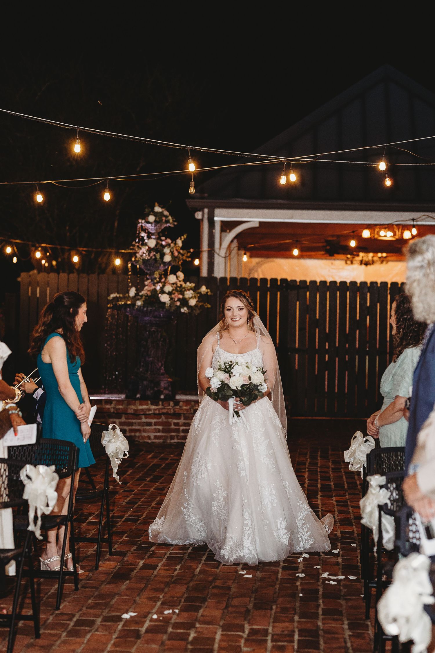 Bride walks down brick aisle under twinkling lights during outdoor evening ceremony.