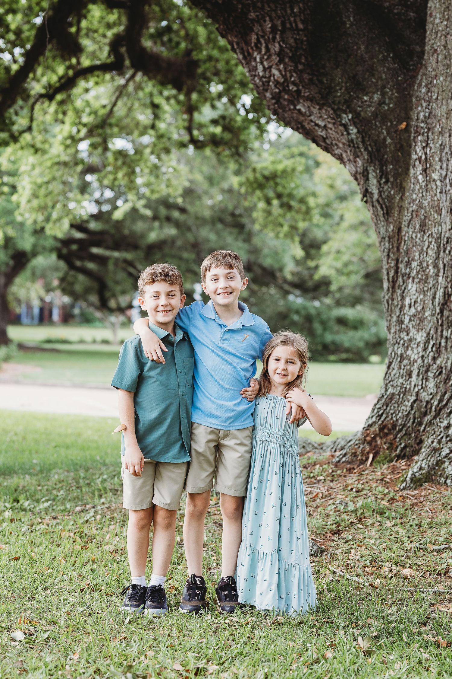 Three children stand together under a large oak tree in a park.