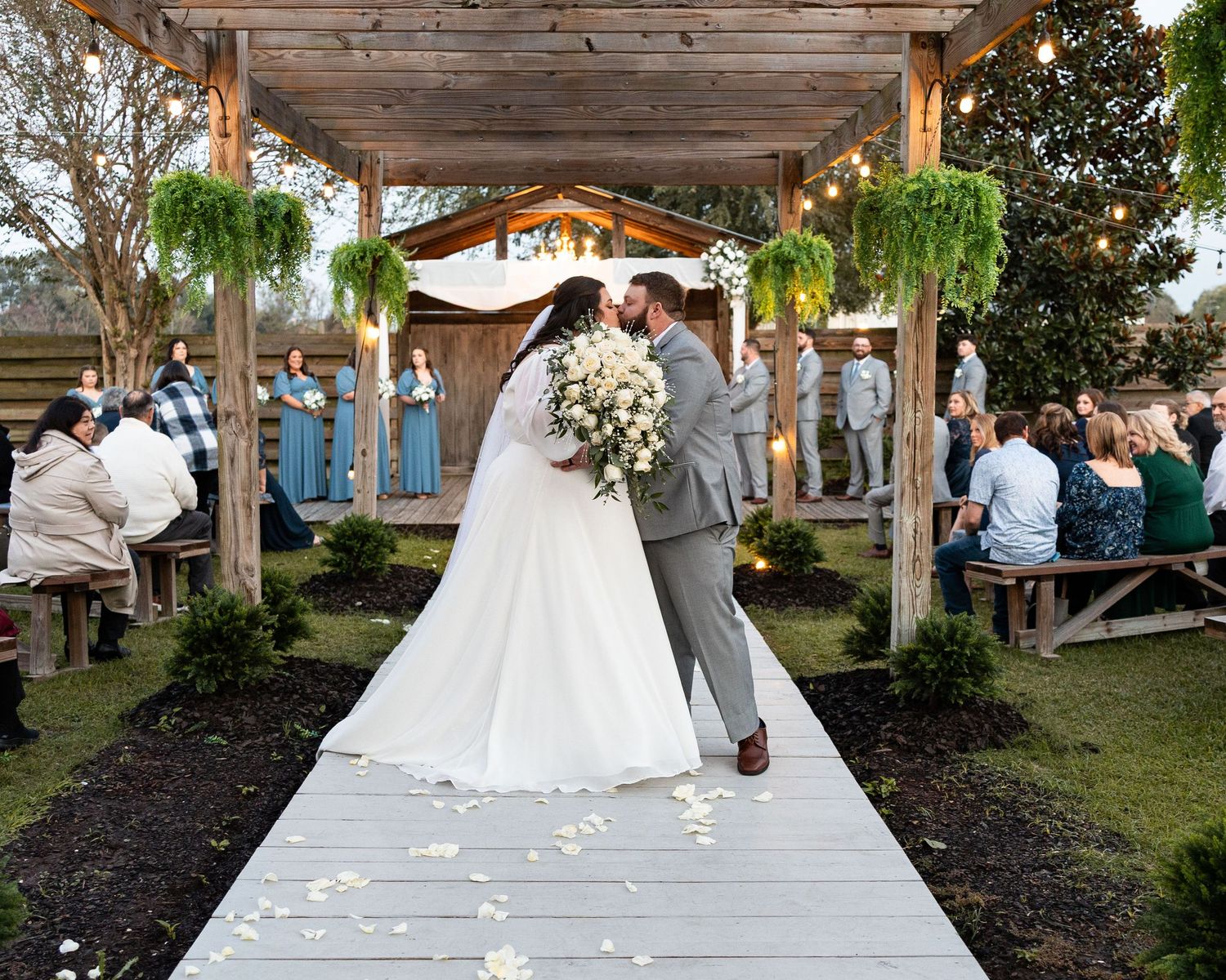 A wedding ceremony procession walks down an outdoor aisle lined with white petals under a wooden pergola.