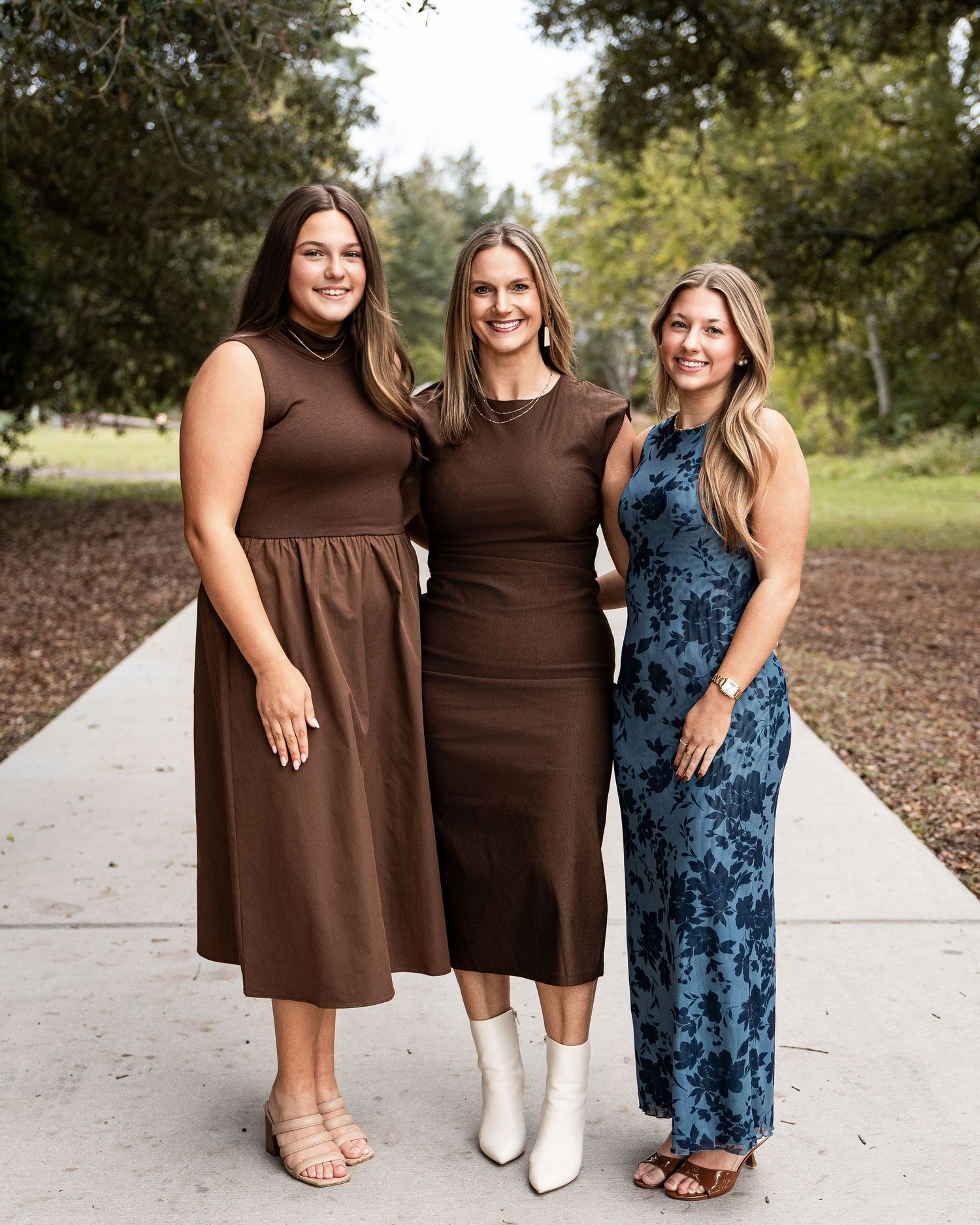 Three people wearing coordinated brown dresses and a blue patterned dress pose together outdoors.