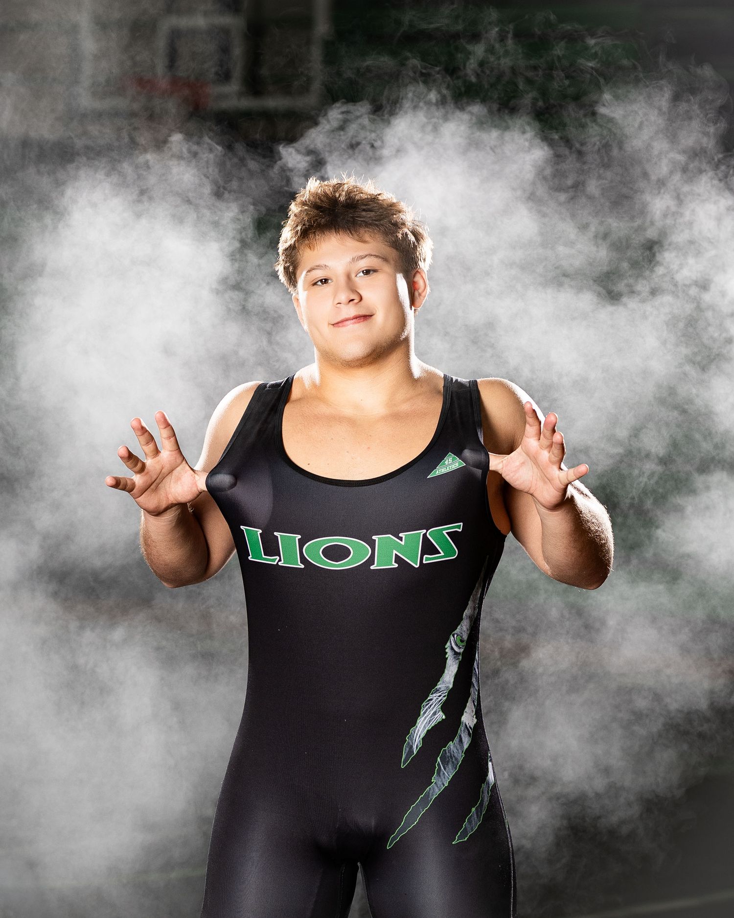High school wrestler in a black Lions singlet poses against dramatic smoky background for sports portrait.