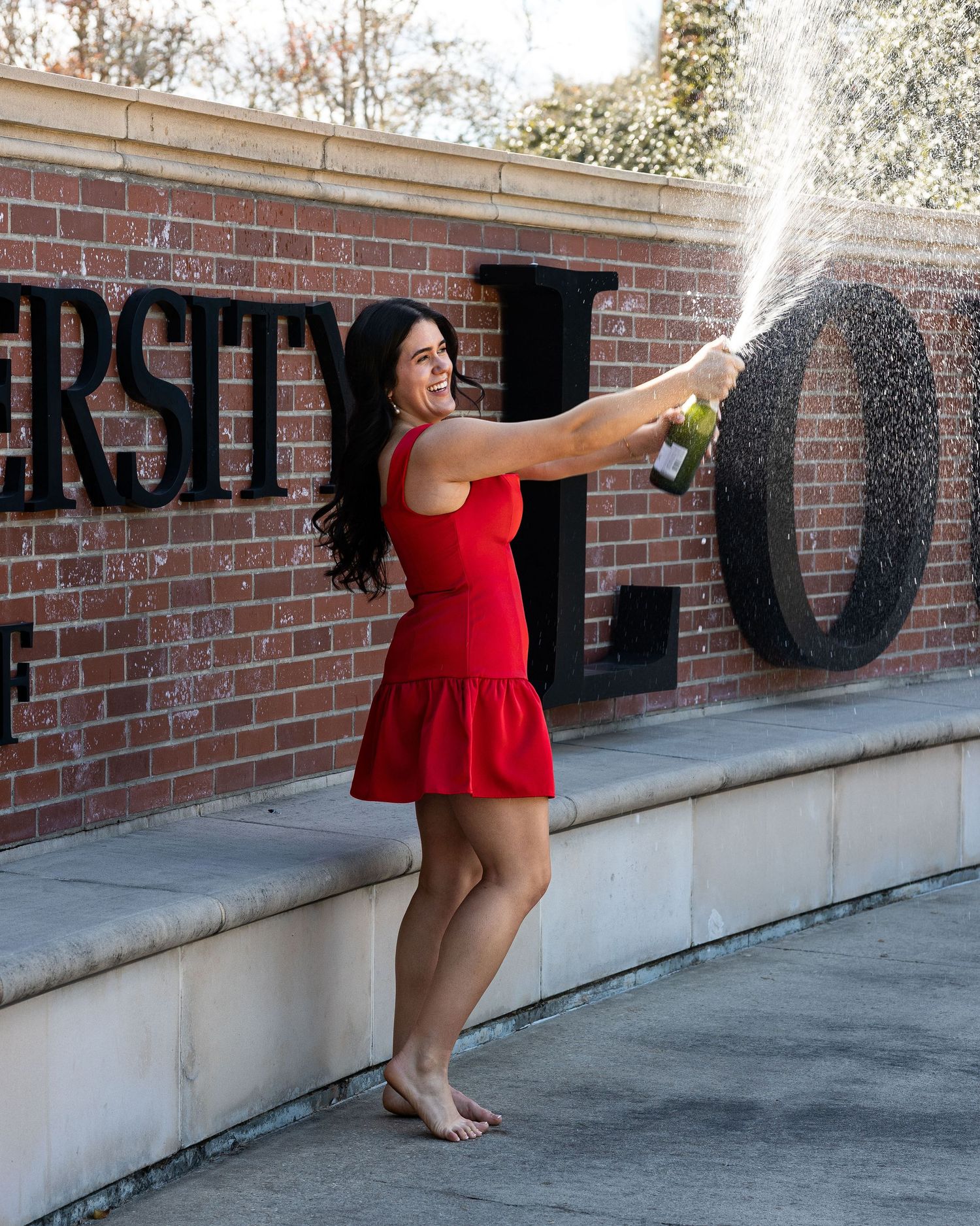 Someone in a red dress sprays champagne while celebrating near a brick wall with lettering outdoors.