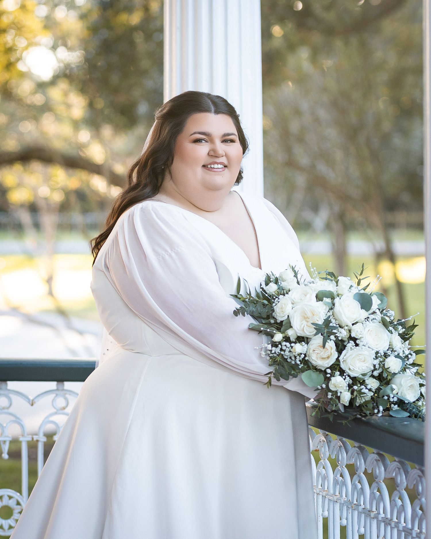 A bride in a white dress holds a white and green floral bouquet while standing on a porch railing in natural light.