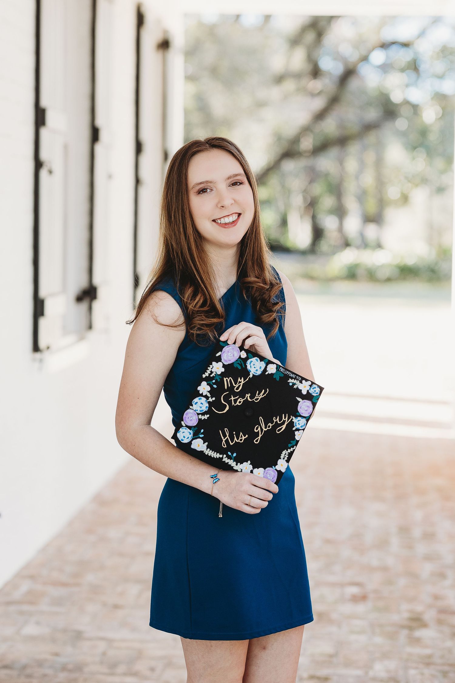 Graduate holding decorated graduation cap wearing navy dress by white columns.