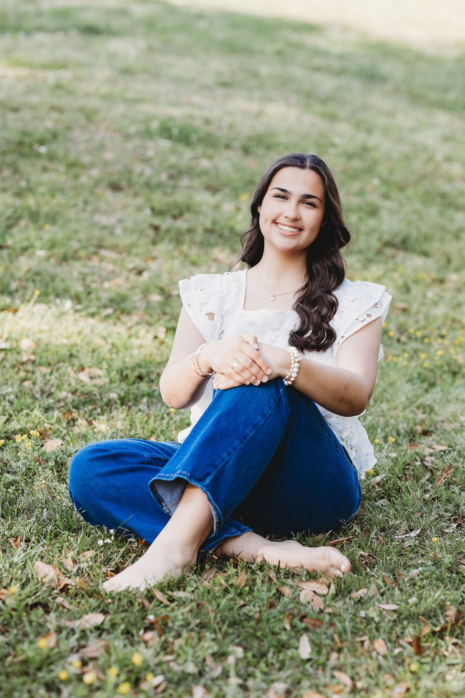 Person in white lace top and jeans sits casually on grass during photoshoot.