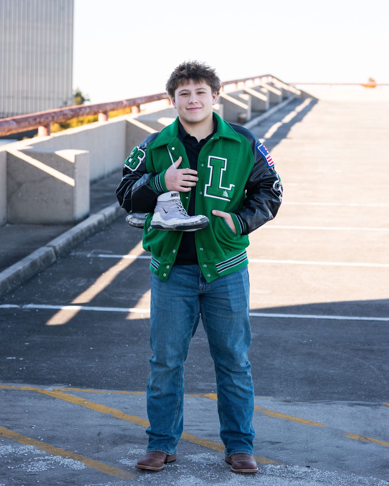 A person in a green varsity jacket and jeans poses playfully in a parking lot on a sunny day.