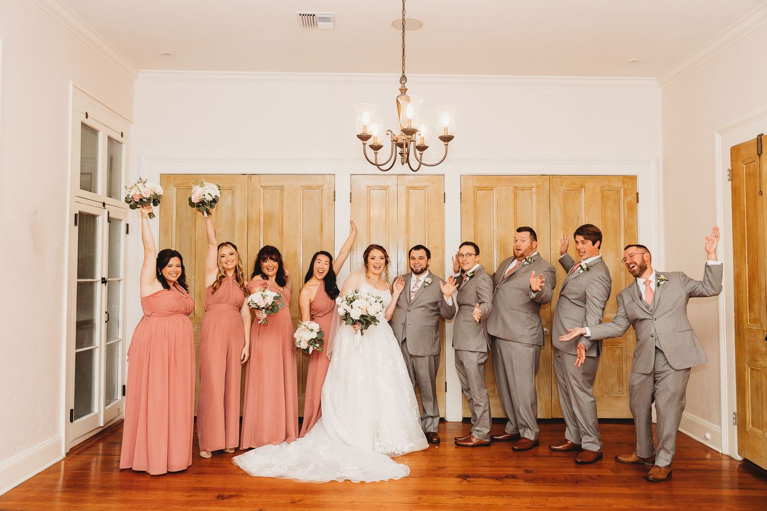 Wedding party strikes a fun pose in coral and grey attire under an elegant chandelier.