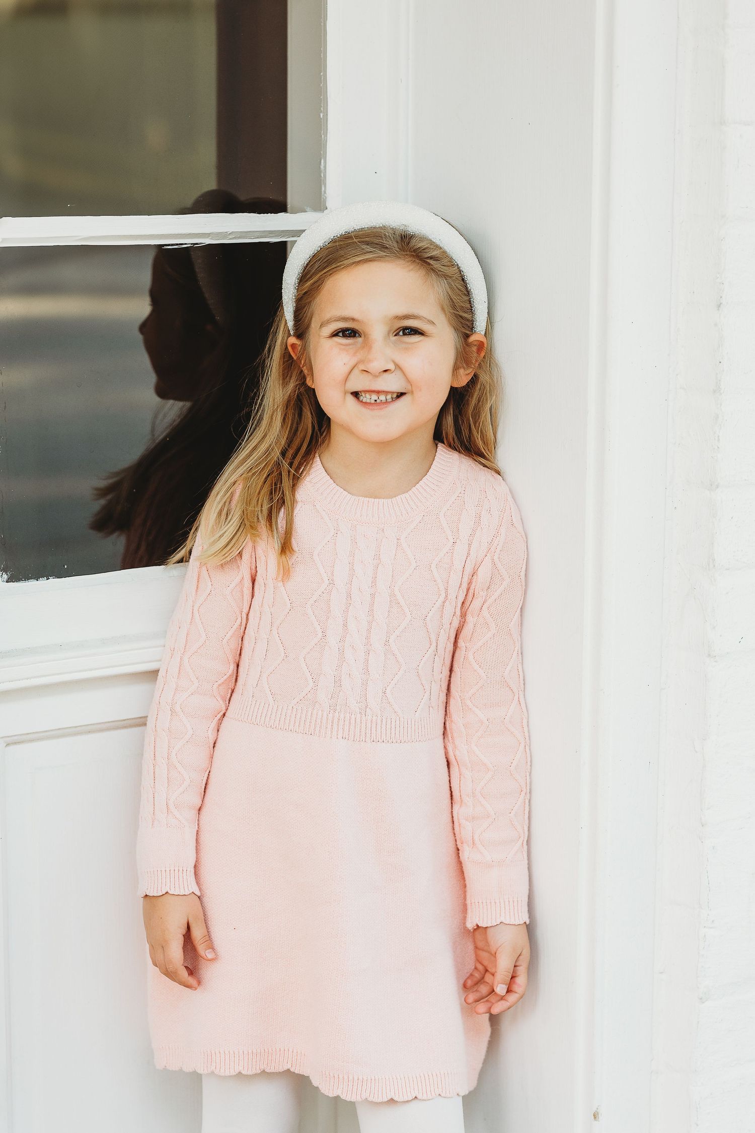 Young person in pink knit sweater poses happily against white architectural backdrop.