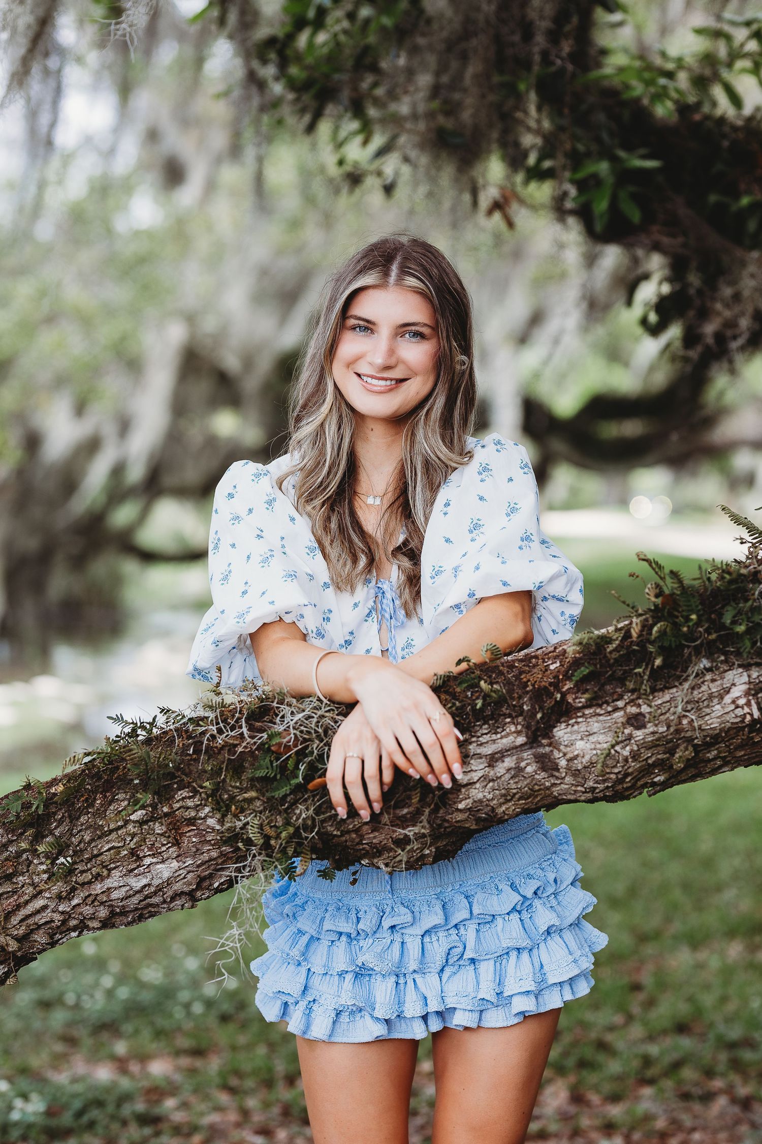 Person in blue floral outfit leaning on tree branch in outdoor setting.