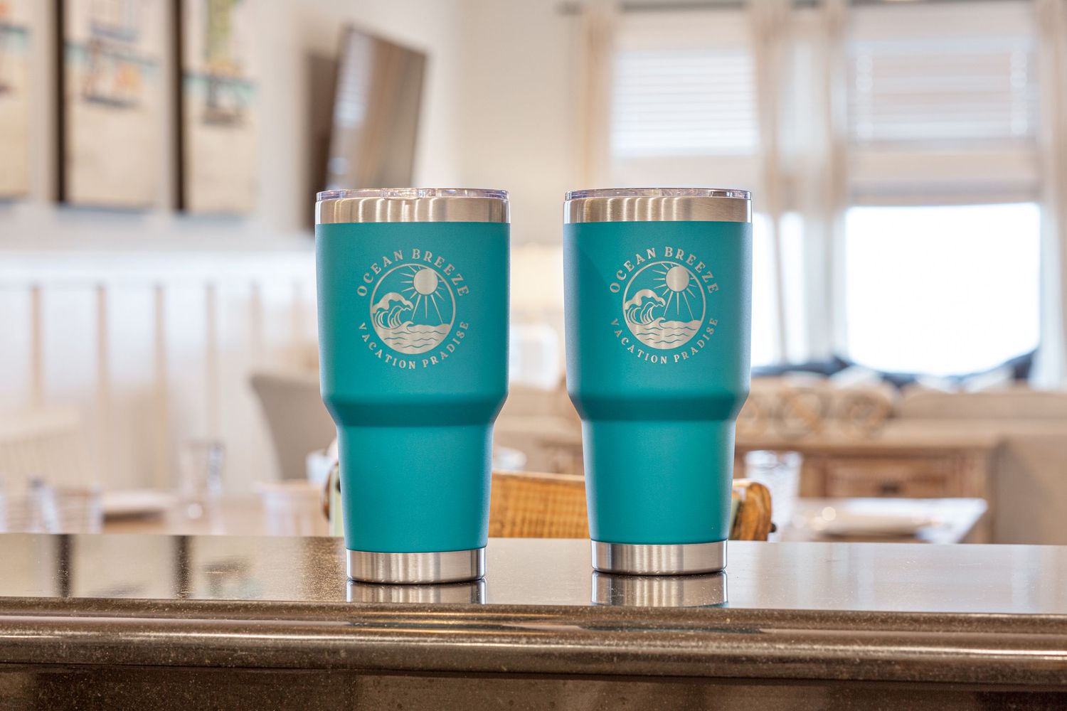 Two teal branded seaside vacation rental tumblers displayed on a wooden countertop indoors.
