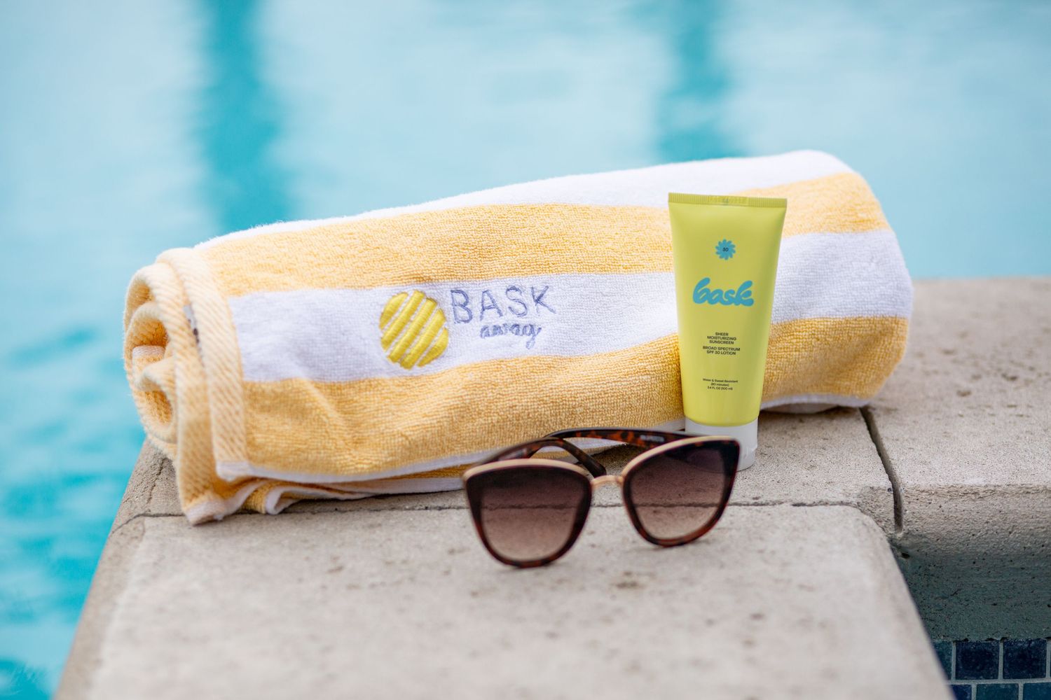 BASK Away branded beach towel, sunscreen, and sunglasses resting on a poolside ledge in summer.