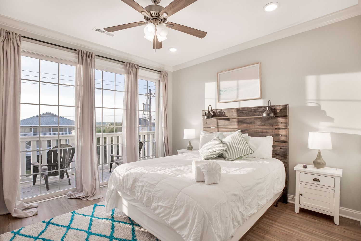Bright coastal bedroom with white bedding, rustic wood headboard, ceiling fan, and balcony access.