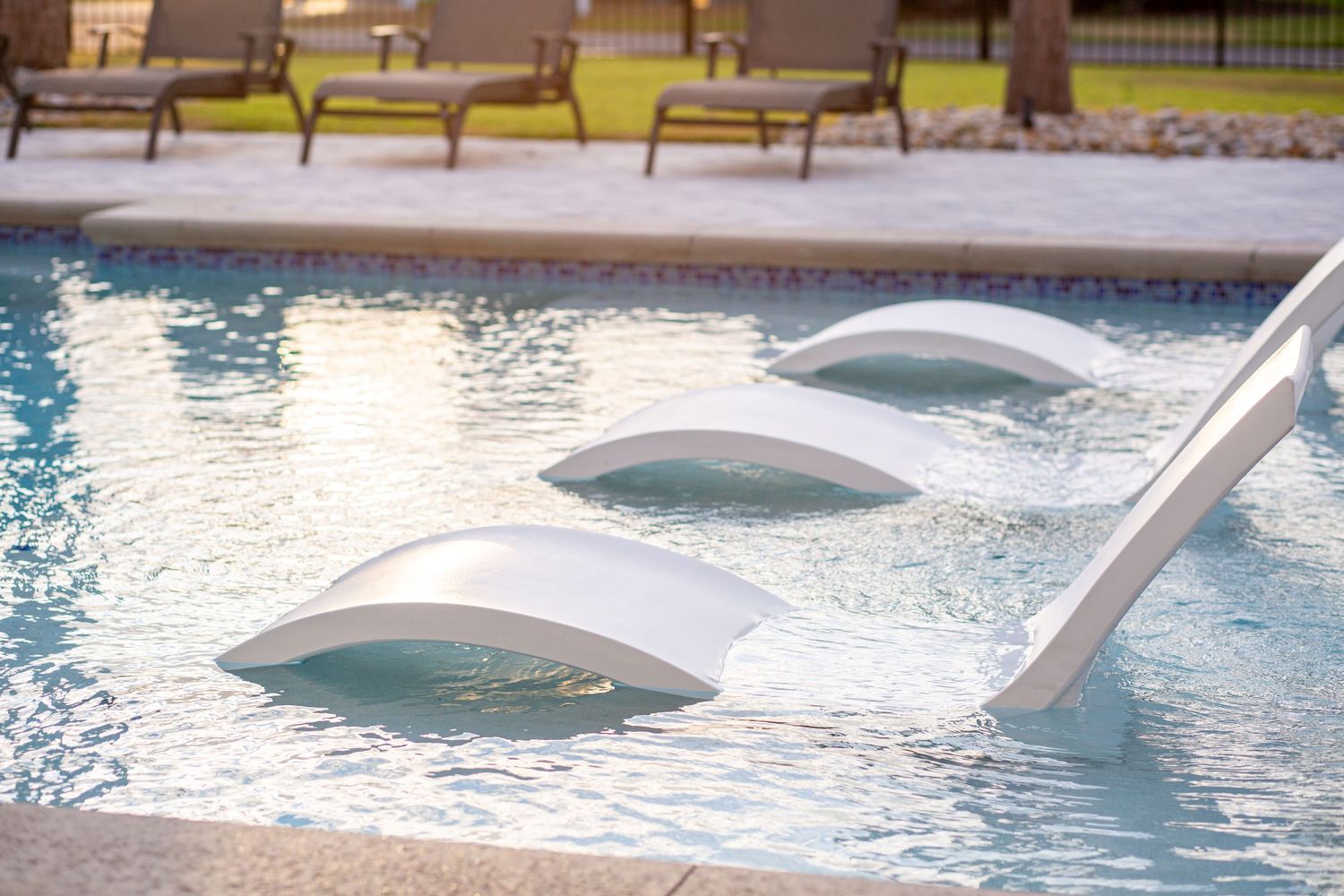 Close-up of white submerged pool loungers partially emerging from crystal clear blue resort swimming pool water.