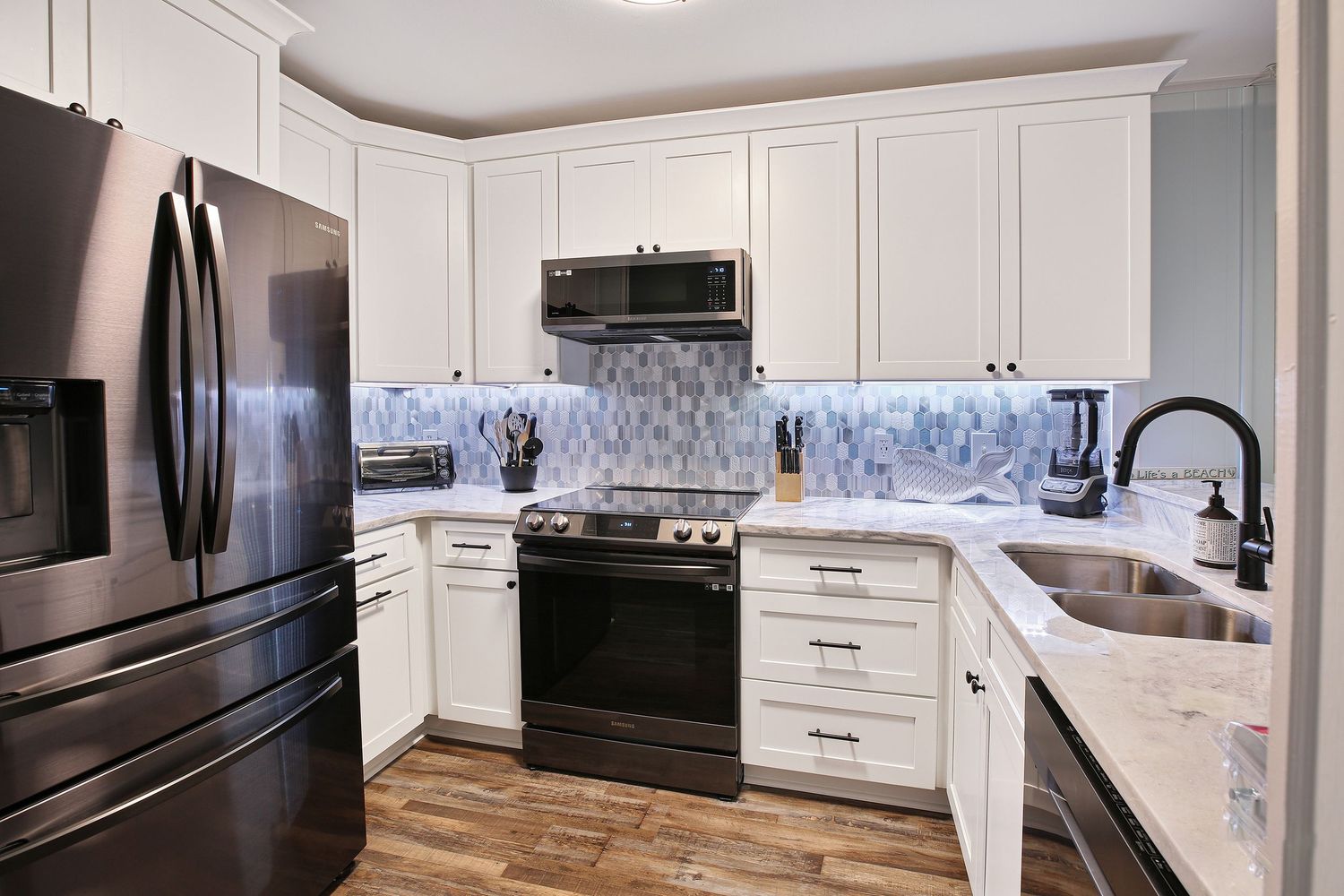 Modern white kitchen with stainless steel appliances, mosaic tile backsplash, quartz countertops, and hardwood floors.