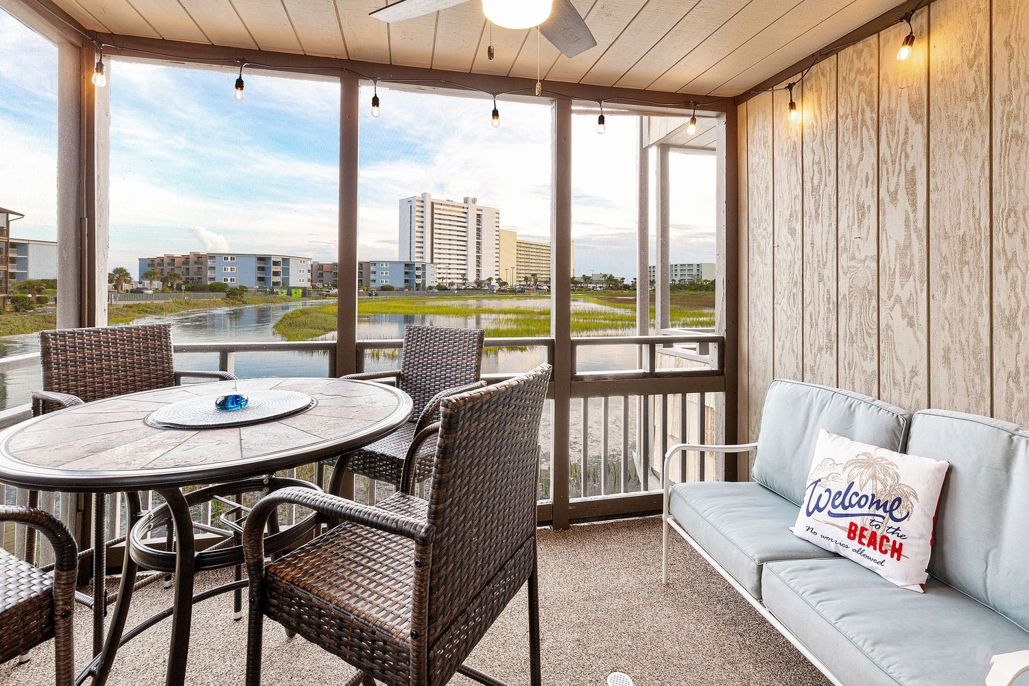 Screened porch with wicker dining set and sofa overlooking a pond and coastal resort buildings.