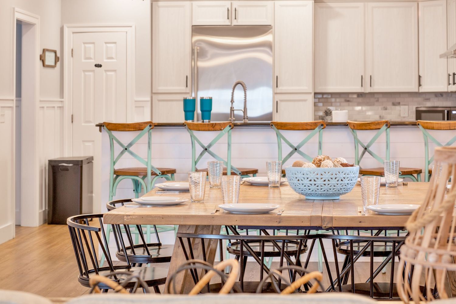 Bright open-plan kitchen and dining area with farmhouse table, wicker chairs, and coastal decor.