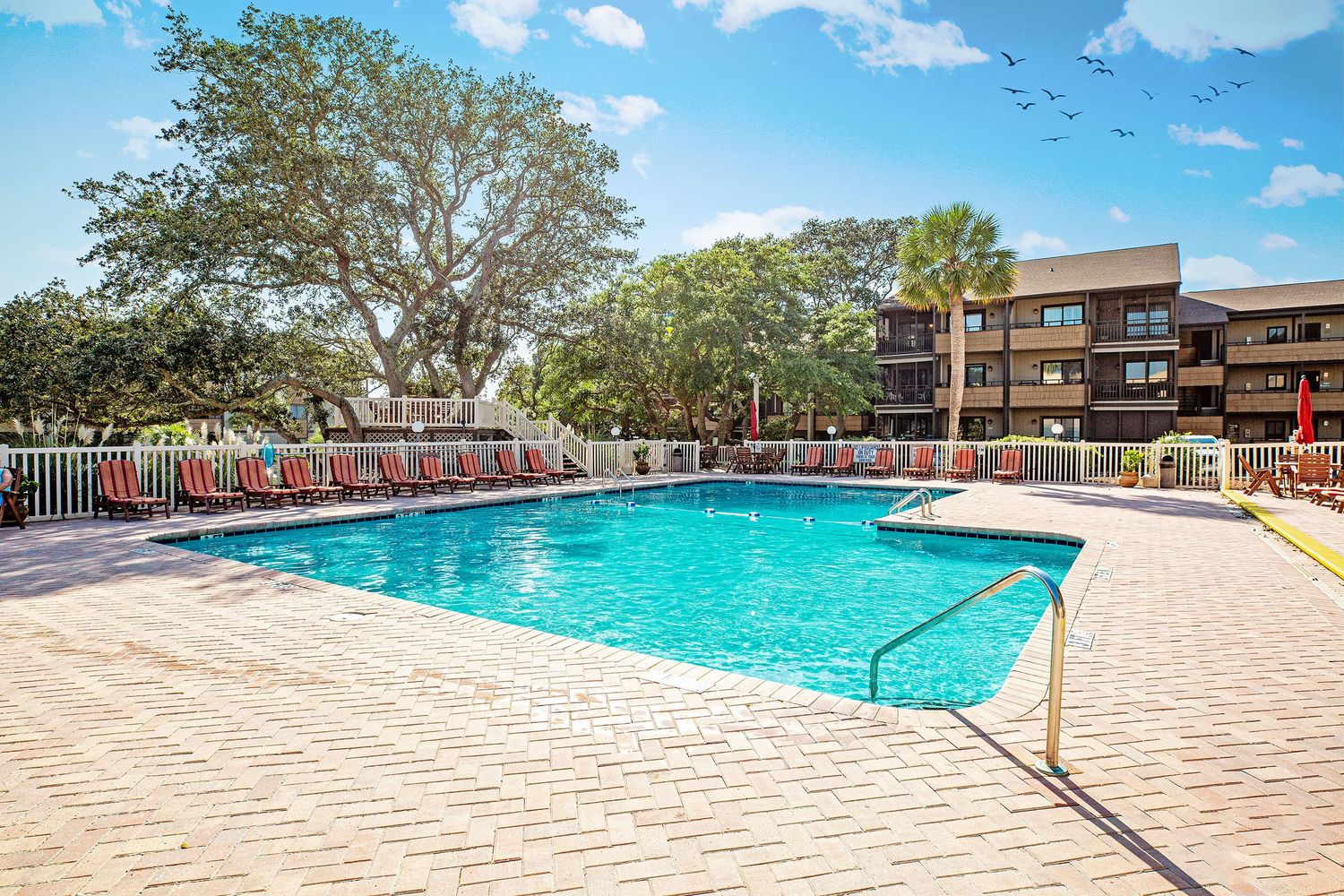 Outdoor community pool surrounded by red lounge chairs, a white fence, and mature trees at Myrtle Beach, near Ocean Annie's.