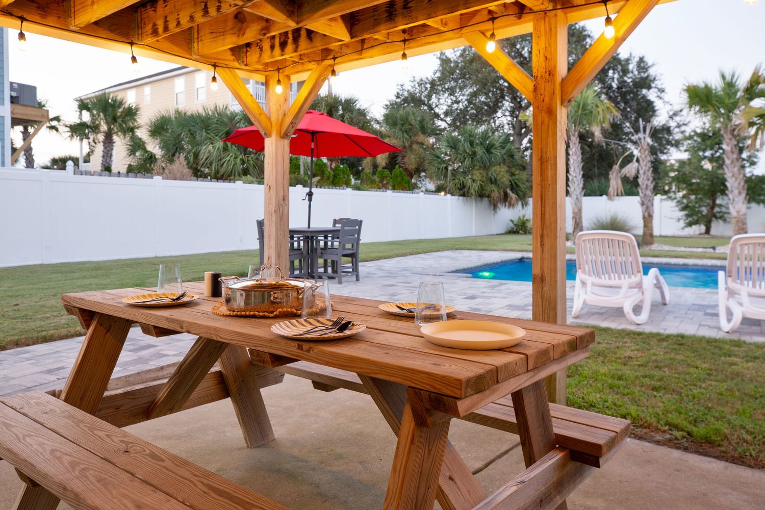 Wooden picnic table set for dining under a pergola beside a sparkling backyard pool.