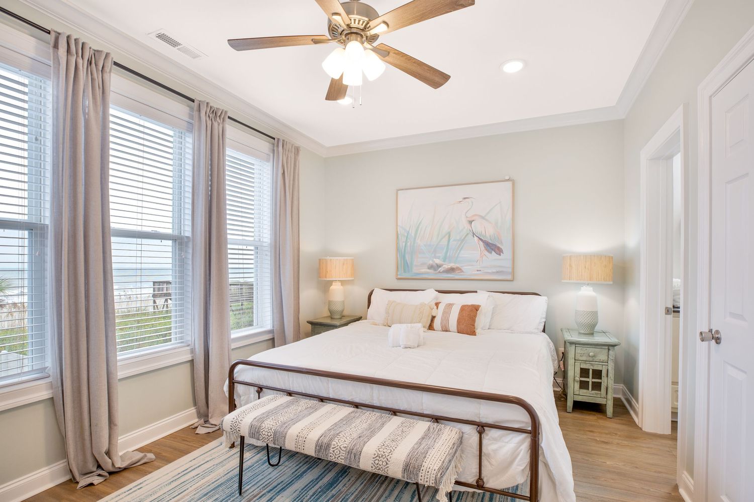 Light and airy coastal bedroom with white bedding, twin nightstands, ceiling fan, and large windows.