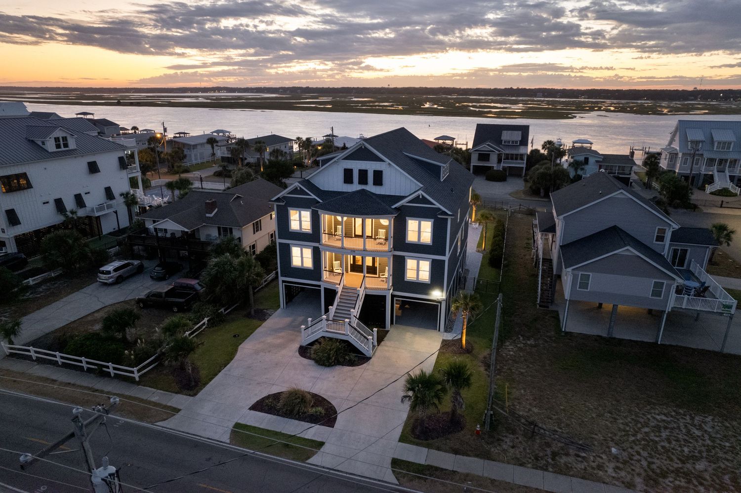 Elegant coastal home glowing at dusk with water views, manicured landscaping, and neighboring beach houses.