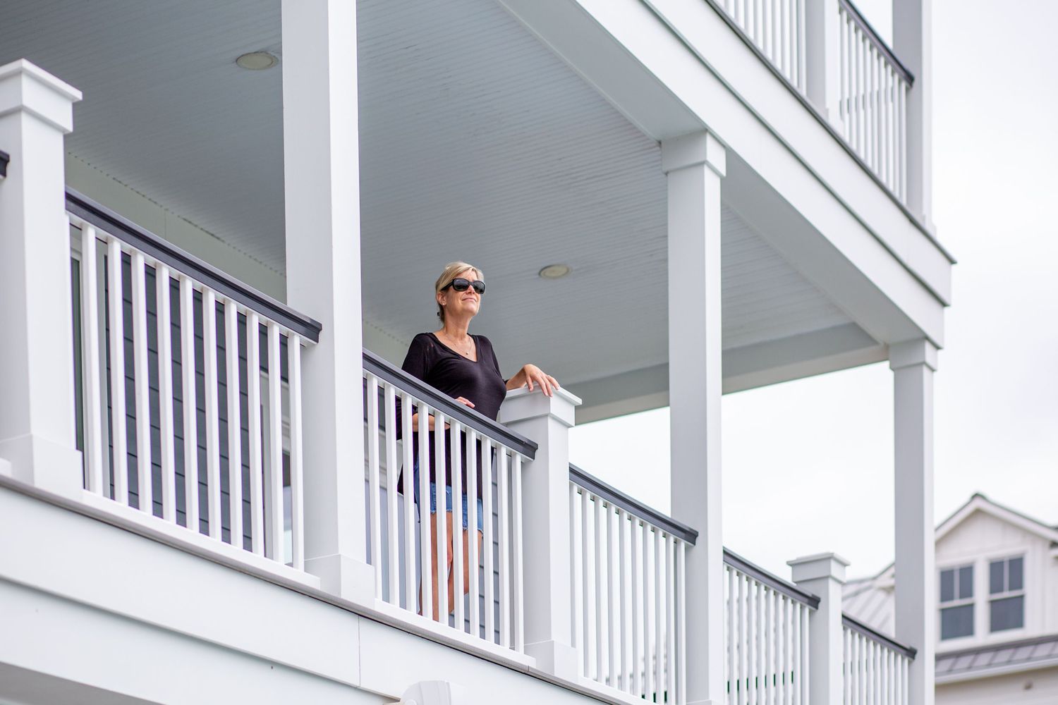 Woman in sunglasses relaxing on a white-railed balcony of a classic coastal beach house.