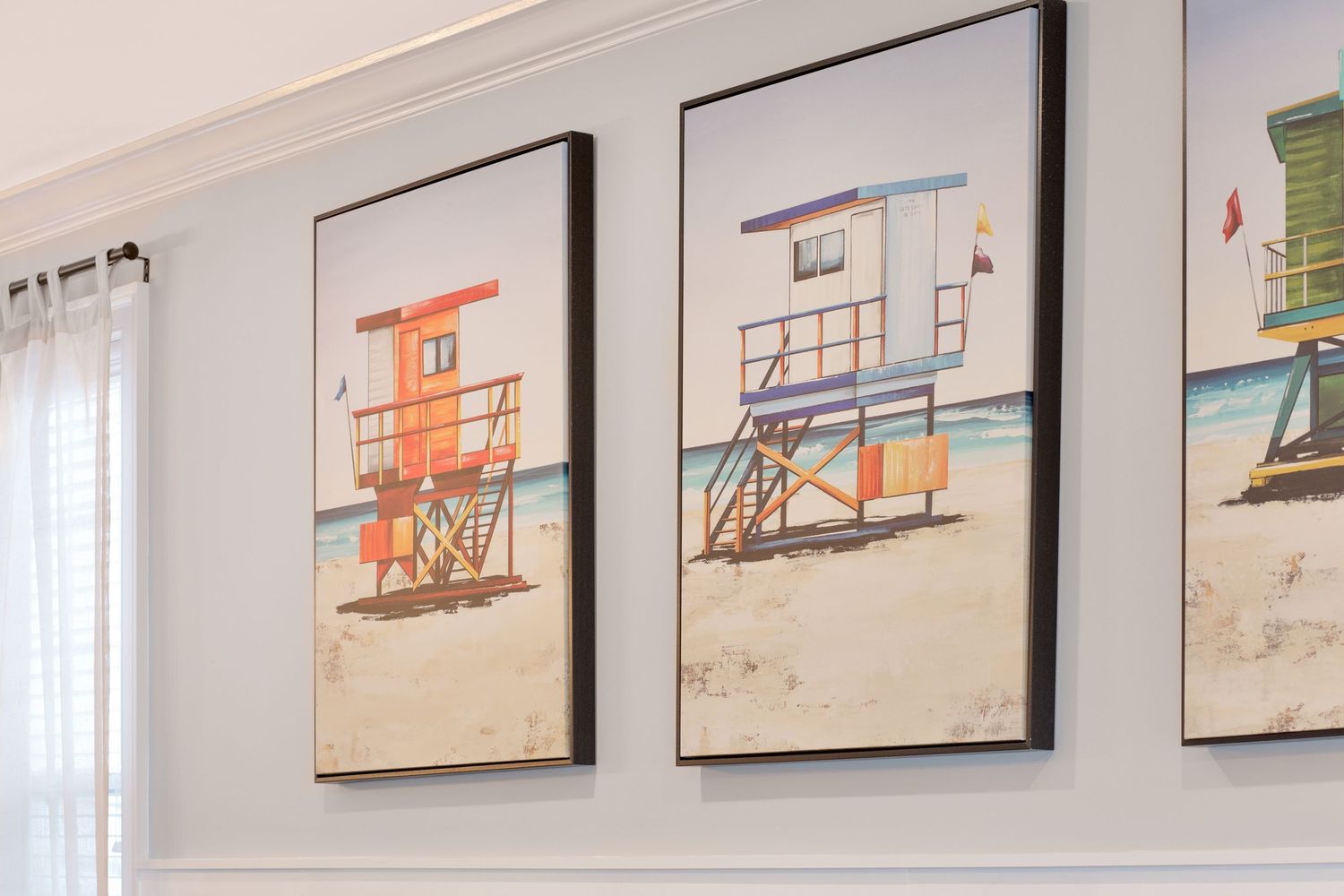 Three framed coastal artwork prints of colorful lifeguard stands on a sandy beach wall display.