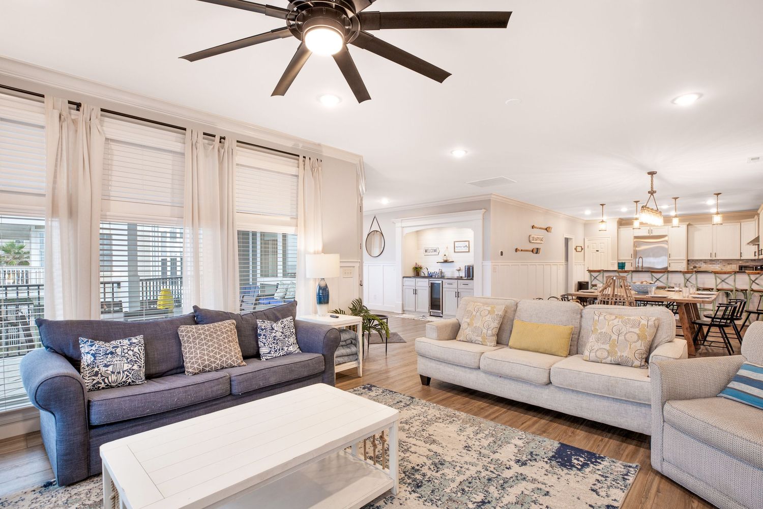 Bright living room with blue and gray sofas, ceiling fan, hardwood floors, and open kitchen.