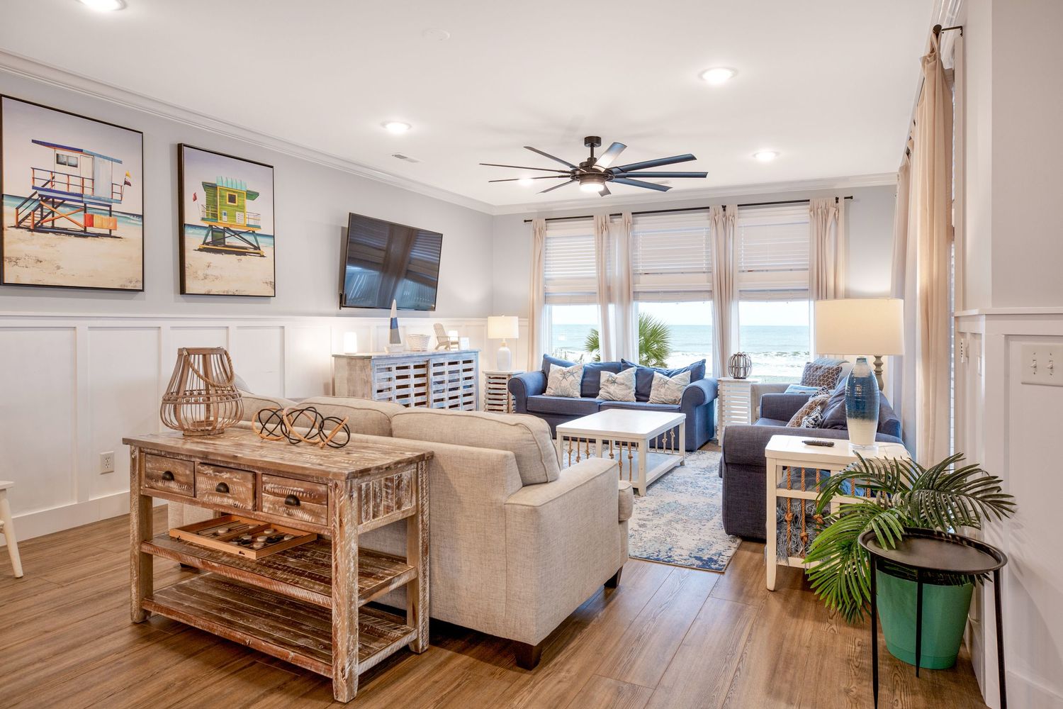 Coastal living room with beige sofas, ceiling fan, wainscoting, and bright natural light windows.
