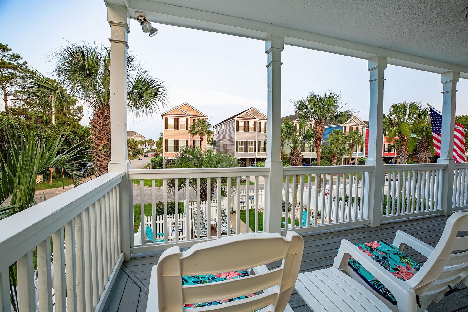 Covered balcony with white chairs overlooking palm trees and colorful coastal neighborhood homes.
