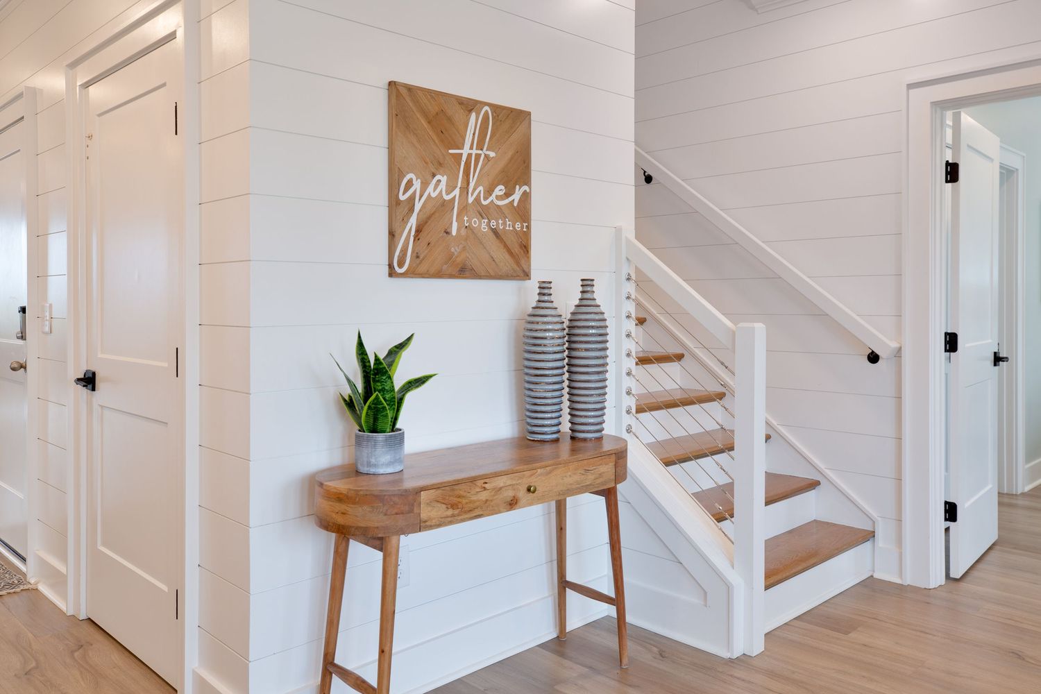 Bright farmhouse entryway with shiplap walls, wooden console table, and a gather sign above the staircase.