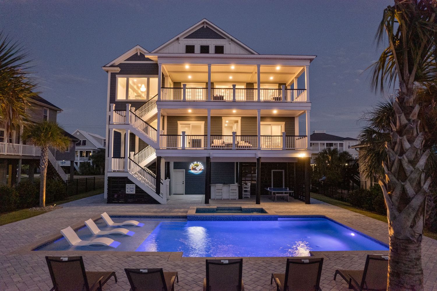 Stunning three-story beach house in Murrells Inlet with lit pool, fire pit, lounge chairs, and palm trees at night.