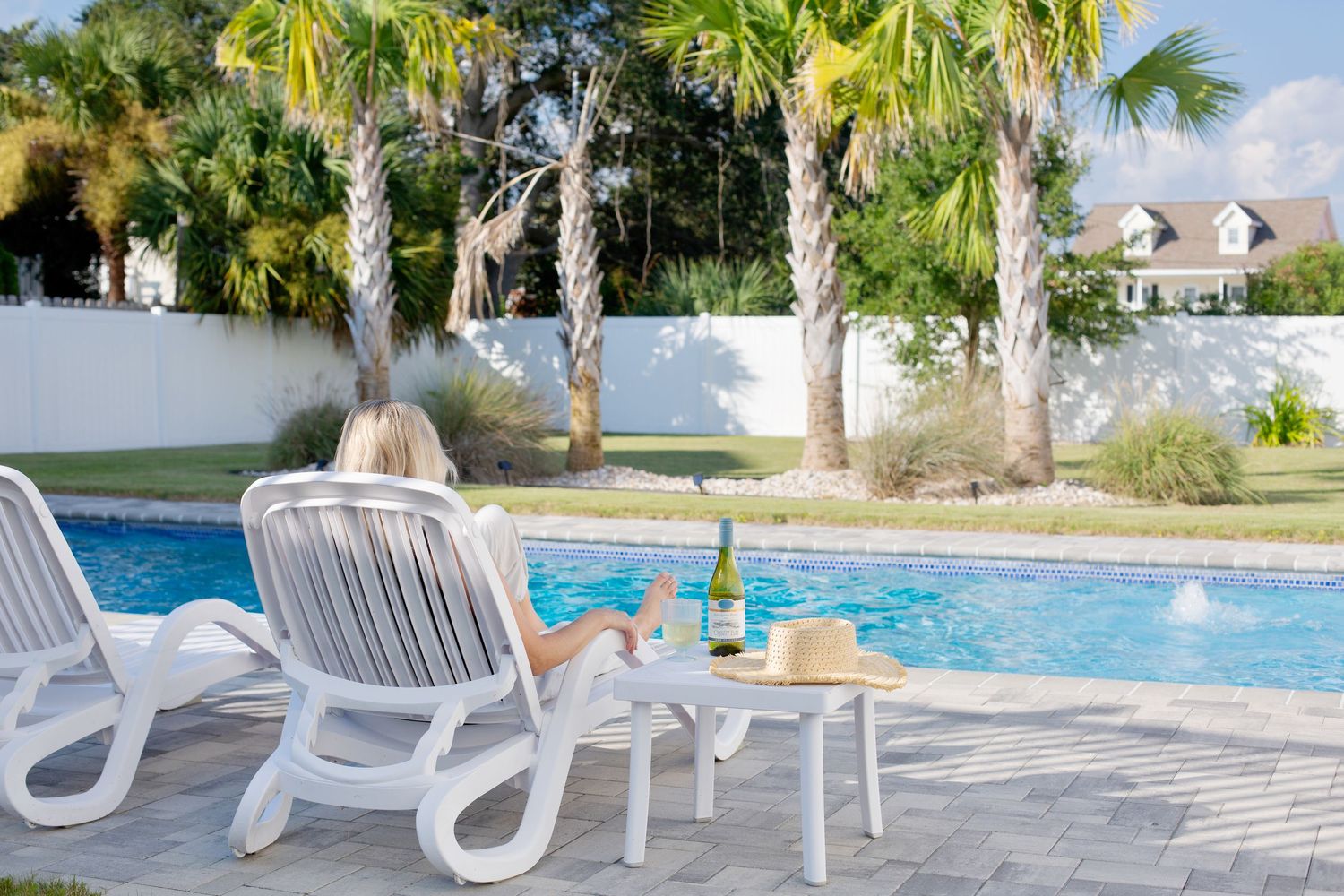 Woman relaxing on lounge chair by a serene pool with palm trees and wine bottle nearby.