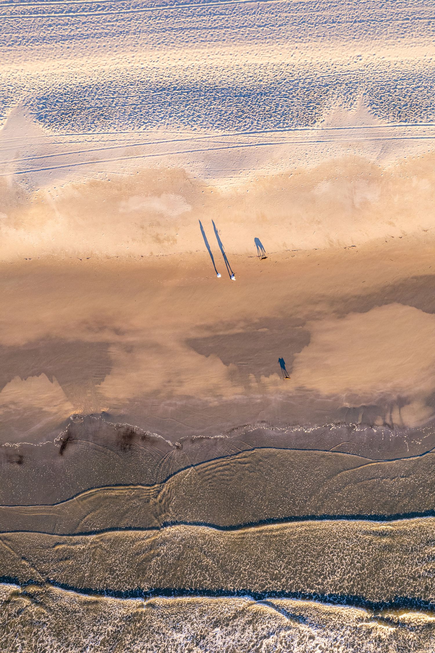 Aerial view of a vast sandy beach with shallow water reflections and two figures casting long shadows. Contact Gabe Rawling, Rawling Photo