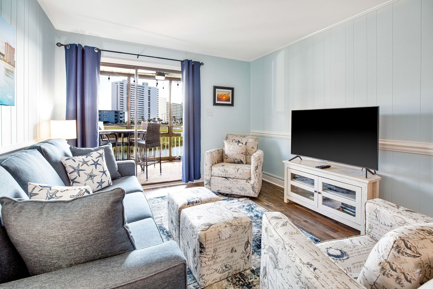 Coastal-themed living room with blue walls, gray sofa, floral armchair, large TV, and sliding doors to a balcony.