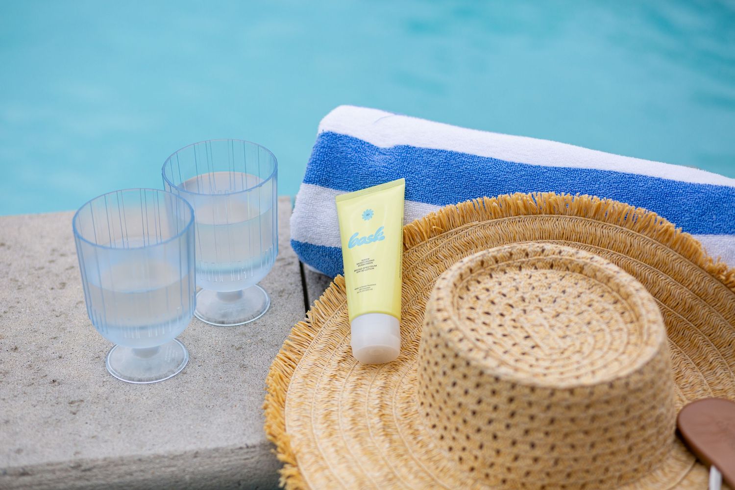 Straw hat, sunscreen, two clear glasses, and striped towel on a sunny poolside ledge. Bask Away Murrells Inlet SC
